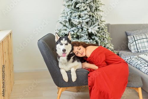 Pretty young girl in red dress siiting on bed and hugs her husky dog over christmas tree