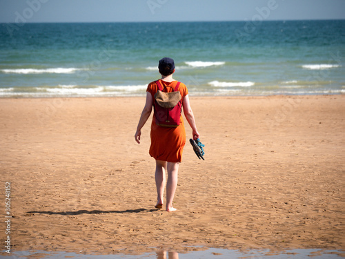 Young woman in oradnge dress walking towards the sea, holding sandals in hand.
