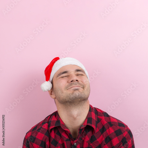 Emotions on the face, smile, joy. A man in a plaid rabbit and a Christmas red hat, on a pink background, copy space.