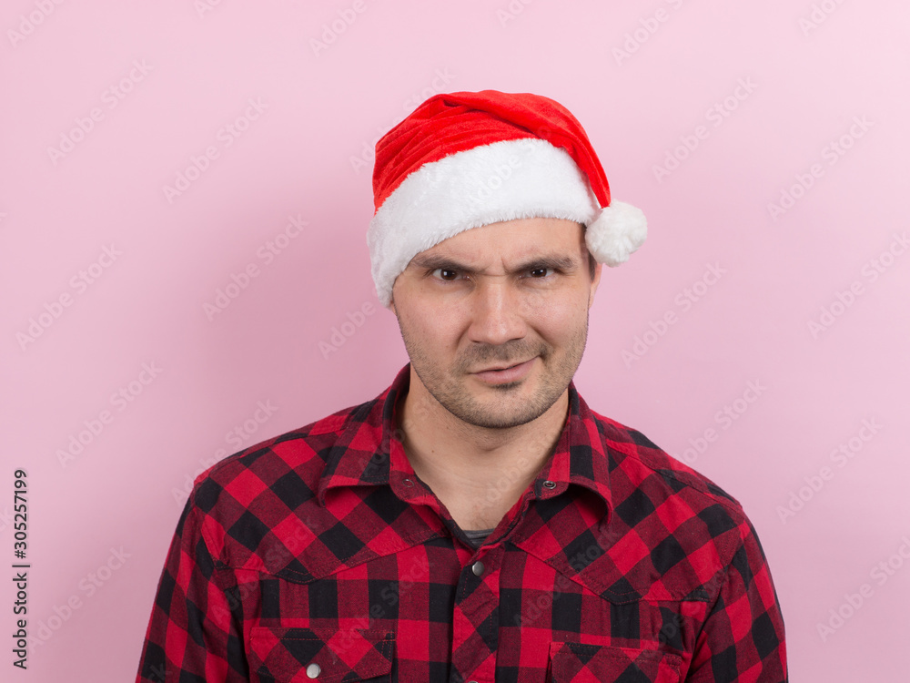 Emotions on the face, angry, bad, bandit, plotting evil. A man in a plaid rabbit and a Christmas red hat, on a pink background, copy space. Emotions on the face, angry, bad, bandit, plotting evil. A man in a plaid rabbit and a Christmas red hat, on a pink background, copy space.