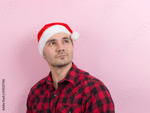 Emotions on the face, pensive, reflection, plan, idea. A man in a plaid rabbit and a Christmas red hat, on a pink background, copy space.