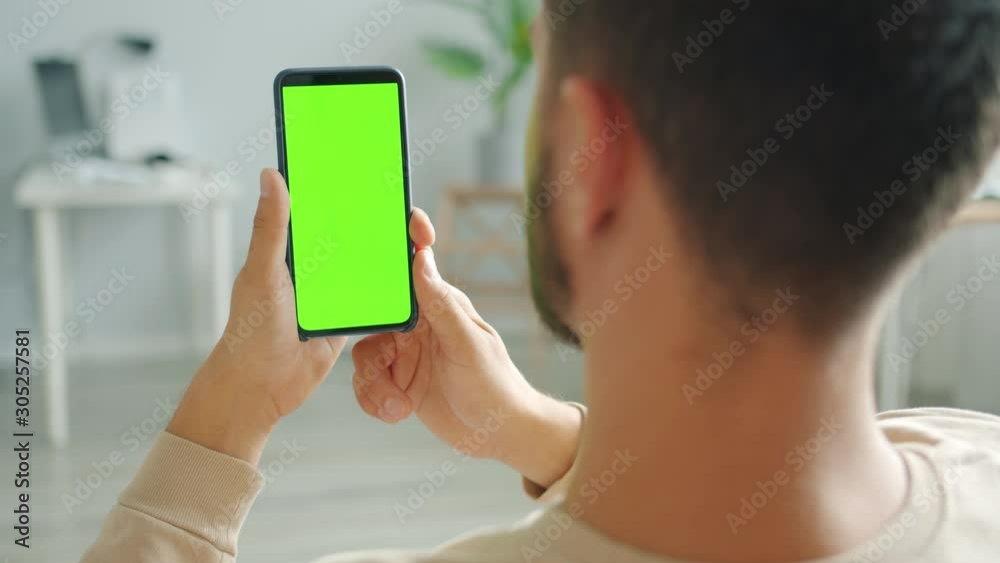 Slow motion of bearded guy brunet is using smartphone with green screen in house holding gadget in hands being in apartment alone. People and devices concept.