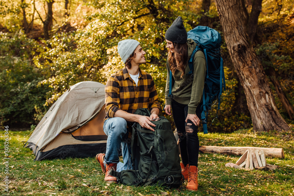 Young beautiful couple gather their backpacks on a hike. Attractive woman and handsome man relaxing together in nature near tent. Young beautiful couple gather their backpacks on a hike. Attractive woman and handsome man relaxing together in nature near tent.