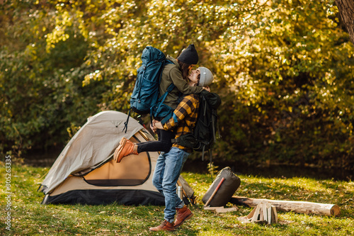 Young beautiful couple with hiking backpack kissing in the forest near tent. Attractive woman and handsome man relaxing together in nature. Instagram mood stories picture. 