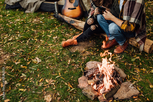 Close up photo of people's legs near tent in camping. Focus on fire. Legs near campfire. Young loving couple of tourists relaxing near the bonfire in the nature.