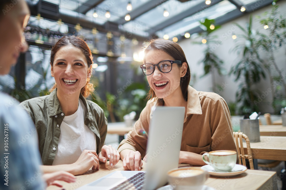 Group of modern young women smiling happily while sitting at table in cafe and using laptop, copy space Group of modern young women smiling happily while sitting at table in cafe and using laptop, copy space