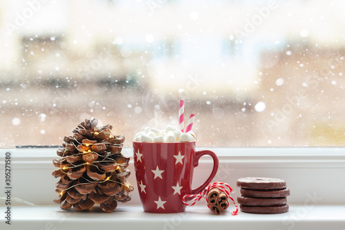 Red cup of hot chocolate drink with marshmallows and cone, cookies, cinamon in front of a window.