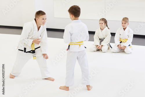 Two boys in kimono standing and practicing technique in karate while other children sitting on the floor and looking at them