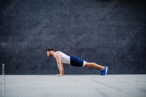 Side view of handsome Caucasian muscular bearded man in shorts and t-shirt doing push-ups. In background is gray wall. © dusanpetkovic1