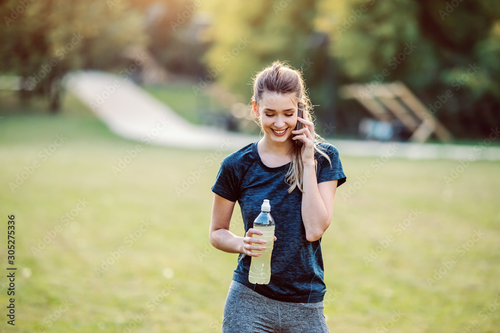 Fototapeta premium Beautiful fit smiling caucasian woman in sportswear and with ponytail standing in nature, talking on the phone and holding bottle with refreshment. Fitness in nature concept.