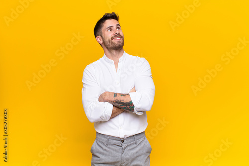 young handosme man feeling happy, proud and hopeful, wondering or thinking, looking up to copy space with crossed arms against flat color wall photo