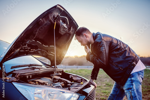 Young serious handsome caucasian bearded blond man in leather jacket looking engine of his car under the hood. Something went wrong.