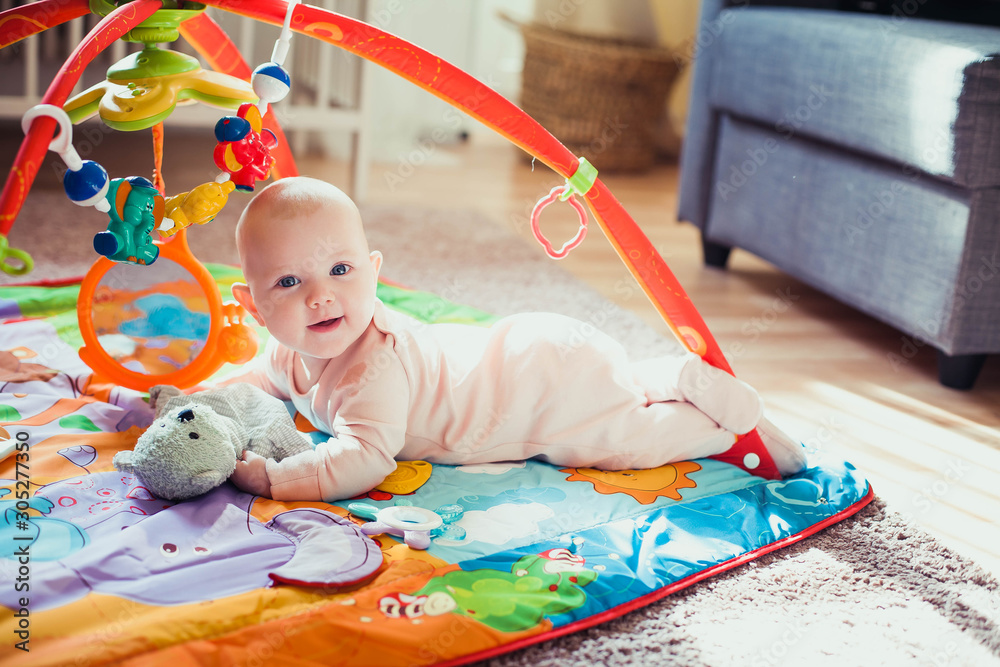 4 months old baby girl lying on colorful play mat on the floor. Activity carpet for kids. - obrazy, fototapety, plakaty 
