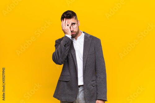 young handosme man feeling bored, frustrated and sleepy after a tiresome, dull and tedious task, holding face with hand against flat color wall