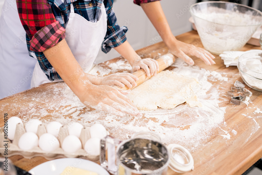 Hands of young female and her son in aprons rolling dough on wooden table Hands of young female and her son in aprons rolling dough on wooden table
