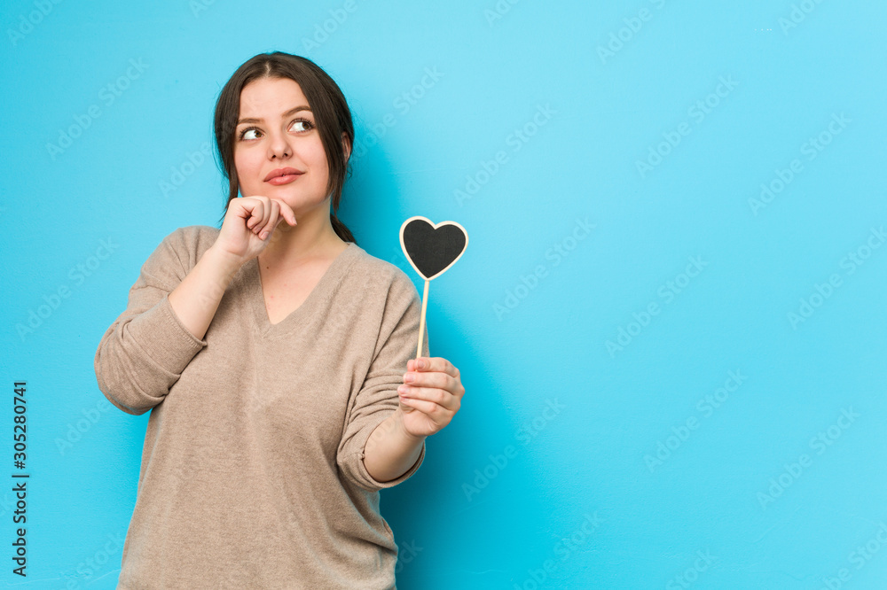 Young plus size curvy woman holding a heart shape looking sideways with doubtful and skeptical expression. Young plus size curvy woman holding a heart shape looking sideways with doubtful and skeptical expression.