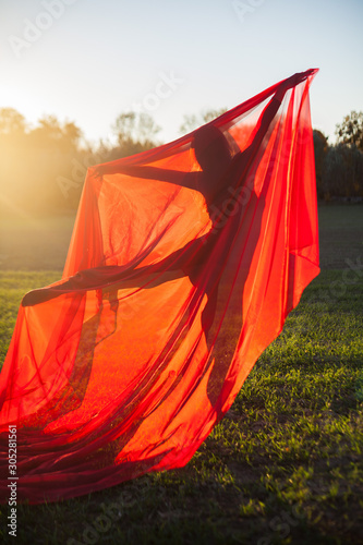 The slender gymnast in black dress in the middle of the field choreographs a large piece of red chiffon in the sunset light.