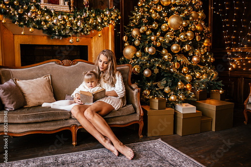 Mother and daughter reading a book at fireplace on Christmas eve. Decorated living room with tree, fire place and gifts. Winter evening at home for parents and kids.
