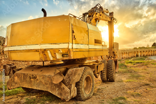 Back view of yellow excavator building in suggestive construction site with Roman aqueduct on background at sunset sunlight. Work in progress, industrial machine. photo