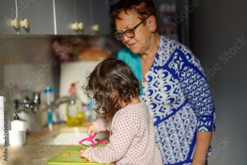 Little cute baby toddler girl with grandmother in the kitchen peeling carrots with carrot peeler on chopping board. Child help at home. Cooking with kids, healthy food, family love.