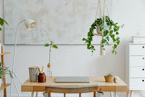 Stylish and boho home interior of living room with wooden desk, laptop, white lamp, macrame shelf and desk supplies. Design and elegant accessories. Modern home decor. Abstract painting on the wall.  photo