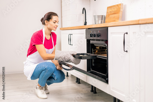 Young housewife squatting while putting tray with raw cookies into electric oven