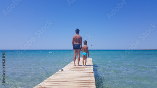 Two brothers on the sea pier. Rear view. Two boys stand on the pier looking into the distance on a Sunny day. Wooden bridge on the beach and blue sky in summer.