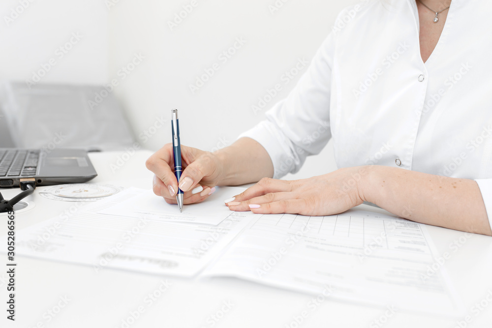 Portrait of a blonde doctor woman in a white coat sitting at a table in a medical office writes notes Portrait of a blonde doctor woman in a white coat sitting at a table in a medical office writes notes