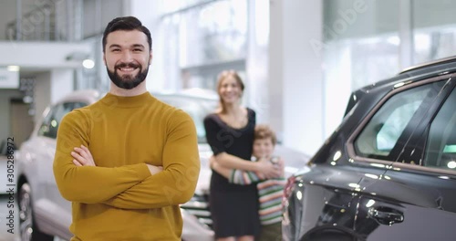 Portrait of satisfied Caucasian man showing car keys, crossing hands, and smiling. His beautiful wife and little son standing at the background next to new automobile. Cinema 4k footage ProRes HQ.