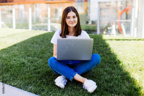 Young woman with laptop sitting on green grass and looking to a display outdoors