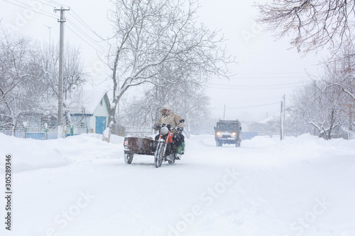 Winter, rural streets are covered with snow. A man rides a motorcycle in a snowstorm