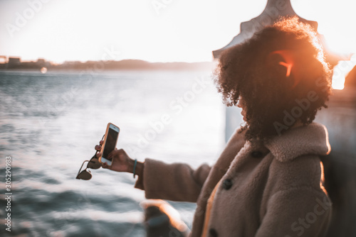 True tilt-shift side view of an African-American girl in a warm demi-season coat taking a selfie with her smartphone in front of the see on a sunny evening, selective focus, shallow depth of field photo