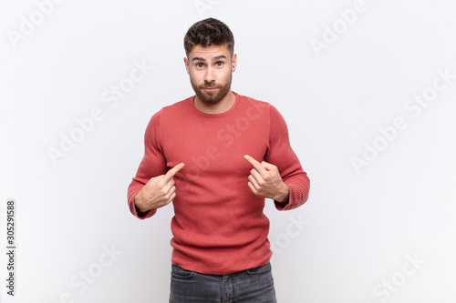 young handosme man pointing to self with a confused and quizzical look, shocked and surprised to be chosen against flat color wall photo