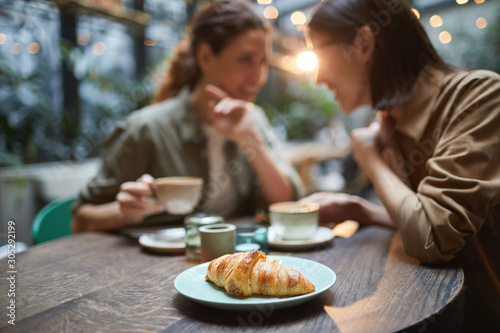 Portrait of two young women gossiping while enjoying lunch together in cafe, focus on fresh croissant in foreground, copy space photo