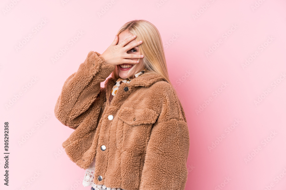 Young blonde woman wearing a coat against a pink background blink at the camera through fingers, embarrassed covering face. Young blonde woman wearing a coat against a pink background blink at the camera through fingers, embarrassed covering face.