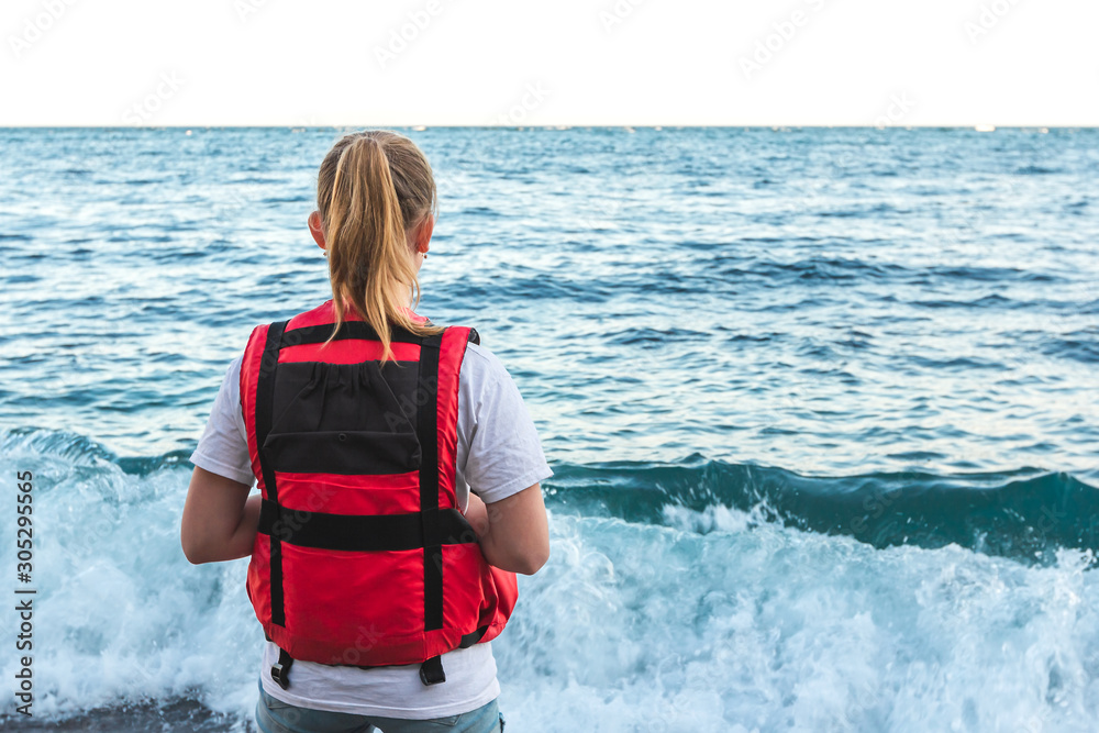 Blonde young girl in life jacket stands on the beach and watching waves.  Lifeguard woman coast