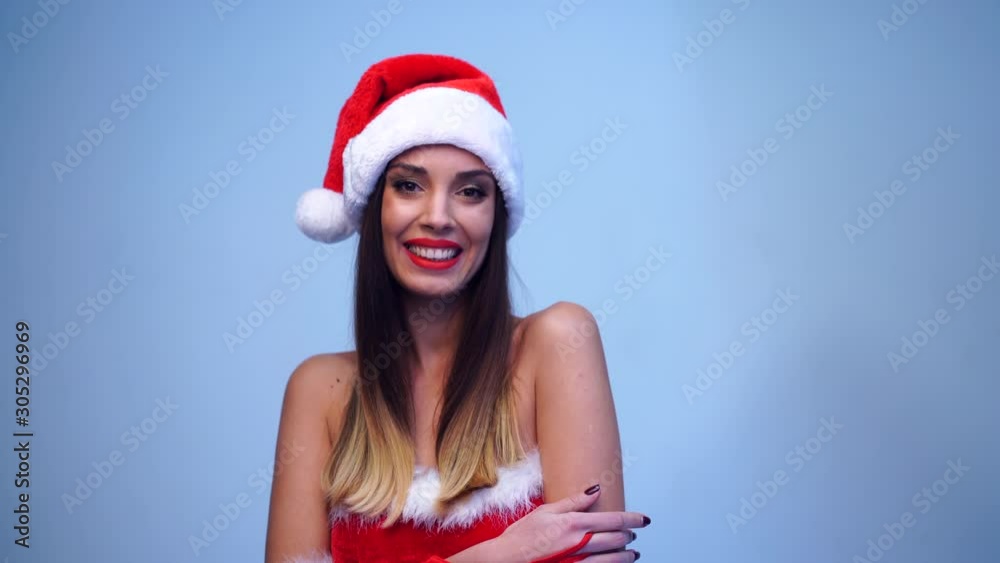 Young beautiful brunette in red dress and Santa Claus hat standing in studio on blue background smiling at the camera Positive Christmas holiday mood Close up portrait Santa girl flirts Young beautiful brunette in red dress and Santa Claus hat standing in studio on blue background smiling at the camera Positive Christmas holiday mood Close up portrait Santa girl flirts