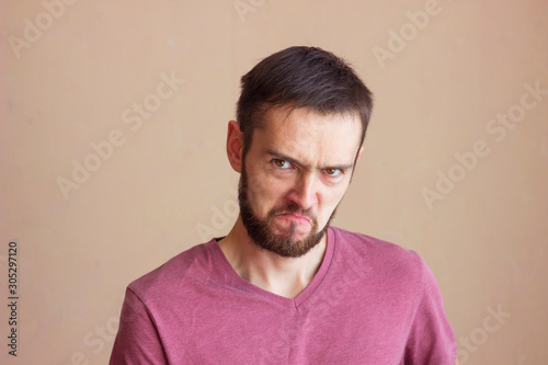 Portrait of young angry bearded man in blank t-shirt, frowning and looks at the camera with disgusted, stands over background. photo