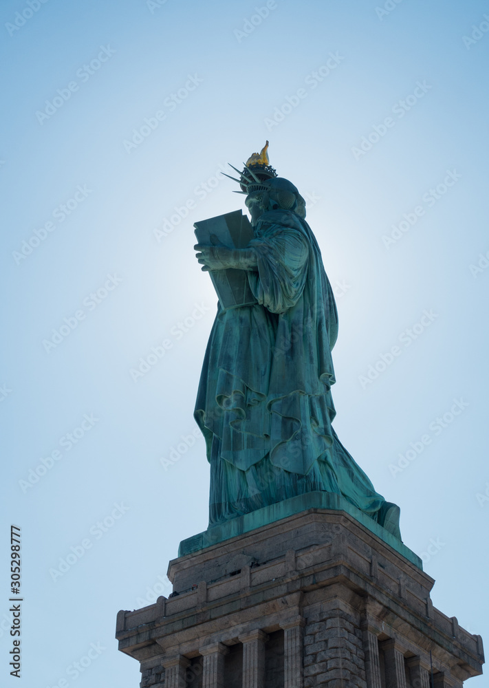 Fototapeta premium Back shot from statue of liberty with sun behind the statue and glowing edges. Torch, Pedestal and blue sky visible.