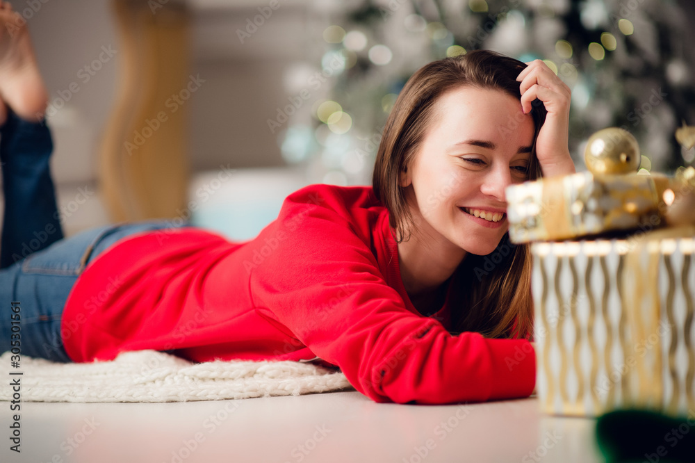 Young smiling woman in sweater holding a gift box celebrating winter holidays in decorated home interior with Christmas tree. Happy New Year concept Young smiling woman in sweater holding a gift box celebrating winter holidays in decorated home interior with Christmas tree. Happy New Year concept