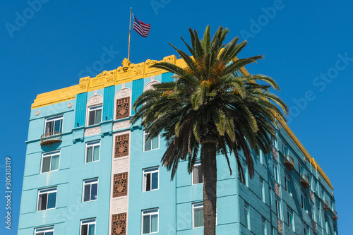 Angled front and side view of a blue art deco style building with yellow trim with an American flag on the rooftop and a palm tree in Santa Monica, California, USA
