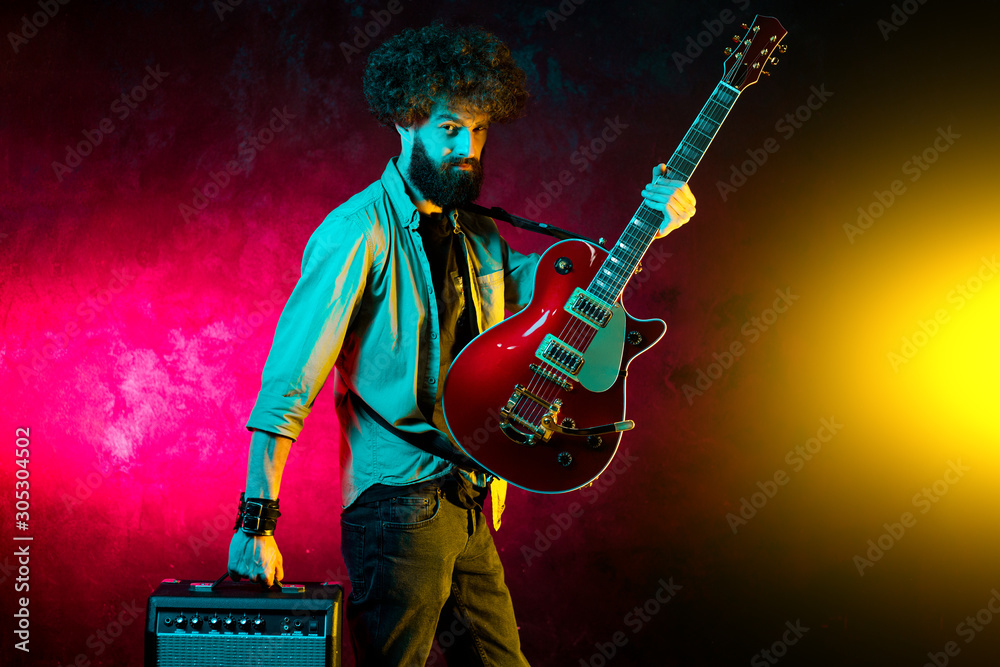 Obraz premium Portrait of hipster man with curly hair with red guitar in neon lights. Rock musician is playing electrical guitar.