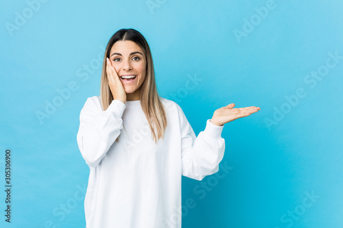 Young caucasian woman isolated holds copy space on a palm, keep hand over cheek. Amazed and delighted.