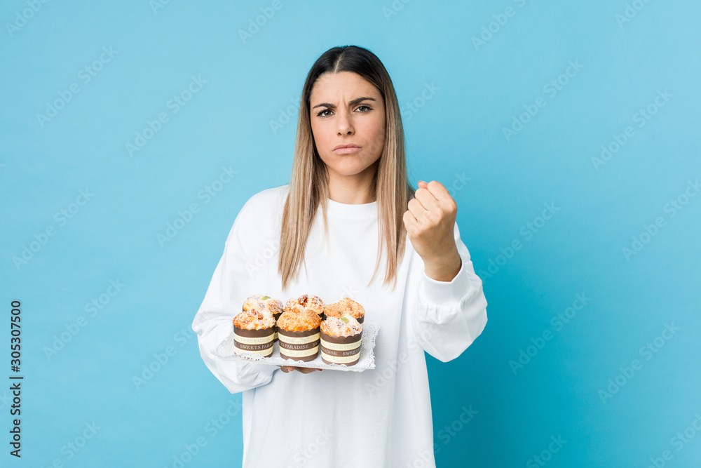 Young caucasian woman holding a sweets cake showing fist to camera, aggressive facial expression. Young caucasian woman holding a sweets cake showing fist to camera, aggressive facial expression.