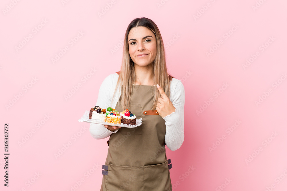 Young baker woman holding sweets pointing with finger at you as if inviting come closer. Young baker woman holding sweets pointing with finger at you as if inviting come closer.