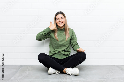 Young caucasian woman sitting on the floor showing a mobile phone call gesture with fingers.