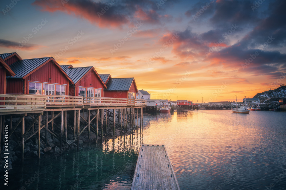 Red rorbu on wooden piles on sea coast, small jetty, colorful sky at amazing sunrise in winter. Lofoten islands, Norway. Traditional norwegian rorbuer, reflection in water, boats in fishing village Red rorbu on wooden piles on sea coast, small jetty, colorful sky at amazing sunrise in winter. Lofoten islands, Norway. Traditional norwegian rorbuer, reflection in water, boats in fishing village