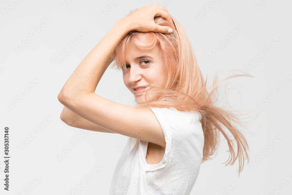 Naklejka premium Studio portrait of cute young attractive blonde girl in white t-shirt. Woman hold hands on head, look at camera and catty smile. Good-looking cheerful female. Youth and beauty concept.