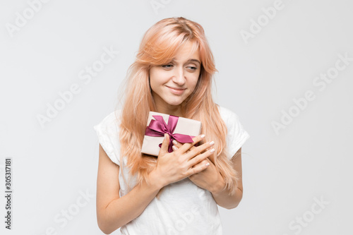 Studio portrait of attractive caucasian girl in white t-shirt who clutches to her chest a gift box with purple ribbon given for Christmas or birthday. Present pleased the girl. Holidays concept.