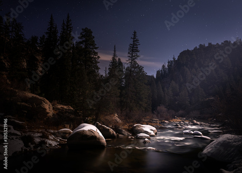 Long exposure of a river at night in the Rocky Mountains. The moon lights the landscape. Stars are visible. The water is smooth. There is a little bit of snow visible. 